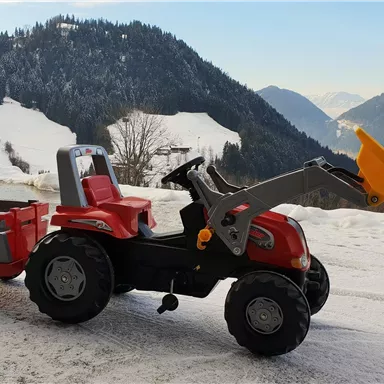 Ein roter Spielzeugtraktor mit einem Anhänger steht auf einem schneebedeckten Gelände. Im Hintergrund sind Berge und ein blauer Himmel zu sehen.