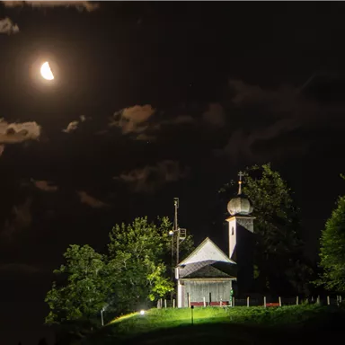 Eine kleine Kapelle steht auf einem Hügel unter einem klaren Nachthimmel. Der Mond leuchtet und Wolken ziehen vorbei.
