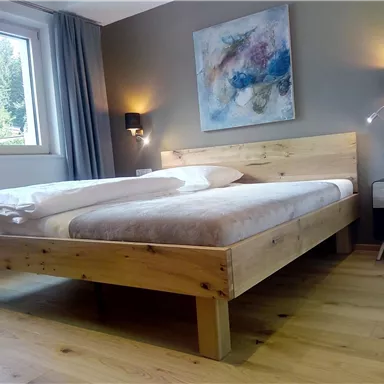 Ein gemütliches Schlafzimmer mit einem Holzbett und modernen Wandlampen. Durch das Fenster ist die grüne Natur sichtbar.