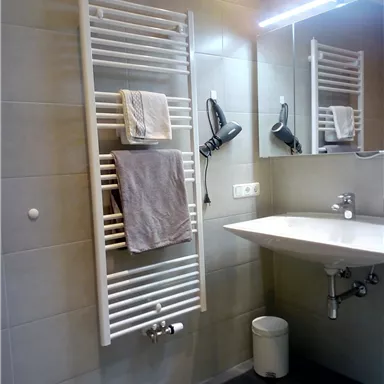 Ein modernes Badezimmer mit einem Handtuchwärmer und einem Waschbecken. An der Wand spiegeln sich die weißen Handtücher und der Badezimmerboden ist grau gefliest.