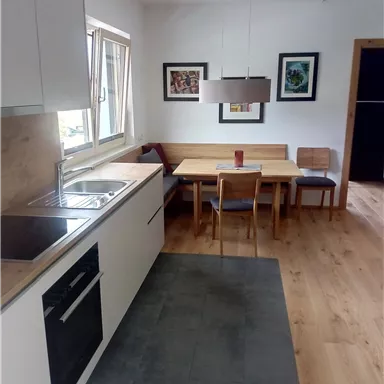 Eine moderne Küche mit hellen Möbeln und einem gemütlichen Essbereich. Die Wände sind mit Kunstwerken dekoriert, und viel natürliches Licht fällt durch das Fenster.