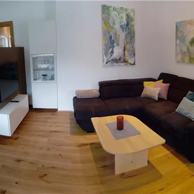 Ein gemütliches Wohnzimmer mit einer großen Couch und einem Kaffeetisch. An den Wänden hängen moderne Kunstwerke und der Boden ist aus hellem Holz.