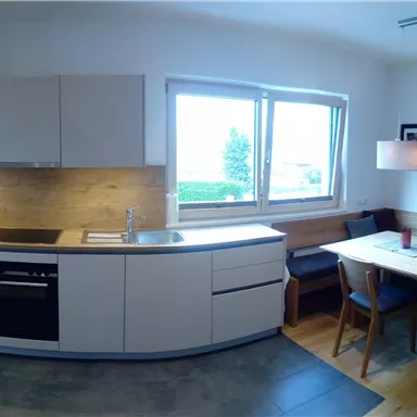 Eine moderne Küche mit eleganten, weißen Schränken und einer hellen Essecke. Große Fenster sorgen für viel Tageslicht.