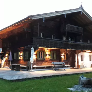 A traditional wooden house in the mountains, illuminated by warm lights. In the background, the mountains and the natural landscape can be seen.