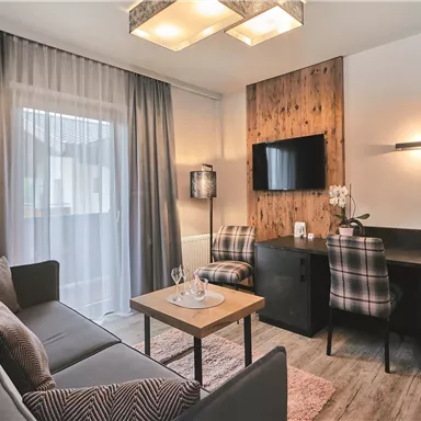 Ein modernes Wohnzimmer mit einem gemütlichen Sofa, einem Tisch und einem Fernseher. Große Fenster sorgen für viel Tageslicht und eine einladende Atmosphäre.