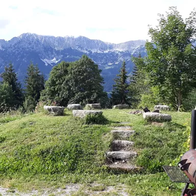 A picturesque landscape overlooking high mountains and a meadow. In the foreground, there are stone blocks and some trees.