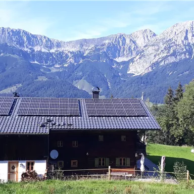 A traditional house with solar panels, surrounded by green meadows. In the background, majestic mountains rise under a blue sky.