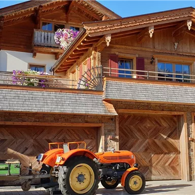 A traditional wooden house with blooming plants at the windows. In front of the house, there is an orange tractor on a paved parking lot.