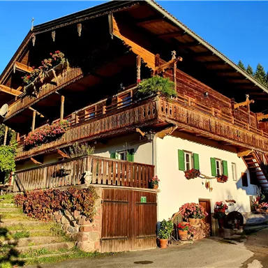 A traditional wooden house with balconies and many flowers. It is located in a rural setting, surrounded by trees.