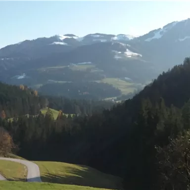 Eine malerische Berglandschaft mit sanften Hügeln und schneebedeckten Gipfeln. Im Vordergrund kurvenreiche Straßen und üppige Wälder.