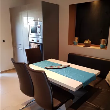 Ein modernes Esszimmer mit einem weißen Tisch und braunen Stühlen. Die Wand ist dunkel, und ein dekoratives Element ist sichtbar.