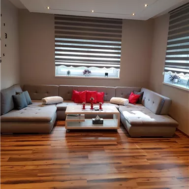 Ein modernes Wohnzimmer mit einem bequemen L-förmigen Sofa und roten Kissen. Große Fenster mit Rollläden und einem hellen Holzfußboden sorgen für eine einladende Atmosphäre.