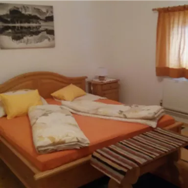 Ein gemütliches Schlafzimmer mit einem Holzbett und orangefarbenen Kissen. Die Fenster sind mit einem hellen Vorhang geschmückt und es gibt einen kleinen Tisch neben dem Bett.
