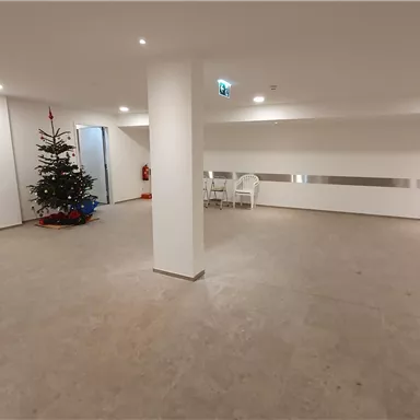An empty room with bright, neutral design. In the corner stands a Christmas tree and some chairs are present.