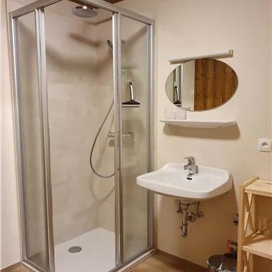 Ein modernes Badezimmer mit einer Glasdusche und einem Waschbecken. Die Wände sind hell und der Boden ist aus Holz.