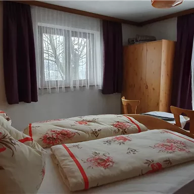 Ein gemütliches Schlafzimmer mit einem Doppelbett und bunten Kissen. Große Fenster lassen viel Licht herein, und im Raum stehen ein Holzschrank und ein Stuhl.