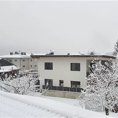 A snowy residential area with white buildings and trees. The sky is gray and the landscape is wintery calm.