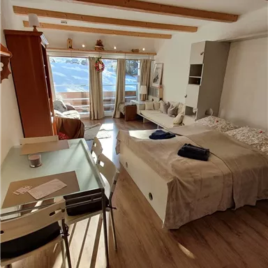 A bright and cozy apartment with wooden ceiling beams. The room includes a bed, a dining table, and large windows with a view.