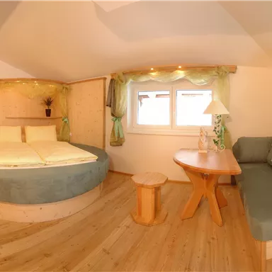 A cozy room with a round bed and large windows. The decor is bright and modern, featuring a small table and a comfortable seating area.