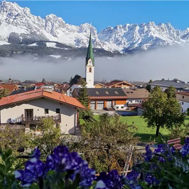 Eine malerische Berglandschaft mit schneebedeckten Gipfeln im Hintergrund. Im Vordergrund sieht man ein kleines Dorf mit einer Kirche und bunten Blumen.