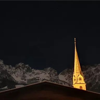 Eine beeindruckende Berglandschaft bei Nacht, mit einer beleuchteten Kirchturmspitze im Vordergrund. Der Himmel ist dunkel und die Berge sind teilweise schneebedeckt.