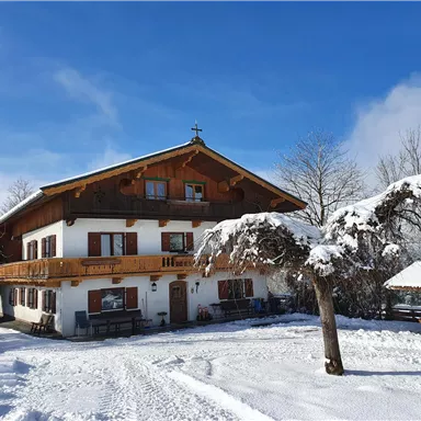 Ein traditionelles Haus im Schnee mit einem schönen Balkon. Umgeben von verschneiten Bäumen und einem klaren blauen Himmel.