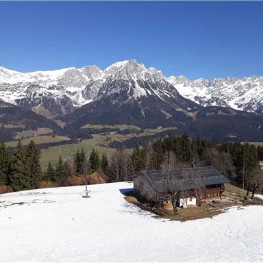 A beautiful alpine landscape with snow-covered mountains. In the foreground stands a typical alpine house on a snow-covered meadow.