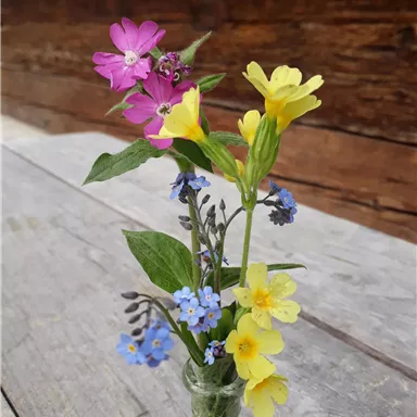 A small glass with colorful flowers in various colors. The flowers are made of yellow, blue, and pink and are on a wooden table.