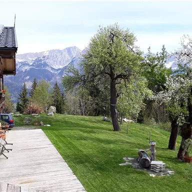 A picturesque garden landscape with blooming trees and a wide view of the mountains. In the foreground, there is a wooden terrace.