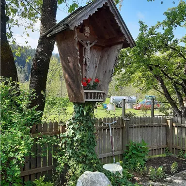 A garden figure in the shape of a wooden chapel with a statue and red flowers. The background shows a green landscape and a fence.