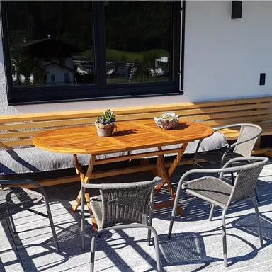 Ein einladender Außenbereich mit einem Holztisch und mehreren Stühlen. Die Fenster bieten einen Blick nach draußen und die Sonne scheint auf den Balkon.
