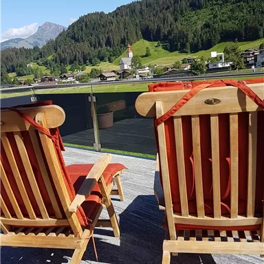 Zwei Liegestühle stehen auf einer Terrasse mit Blick auf grüne Hügel und Wälder. Der Himmel ist klar und blau.