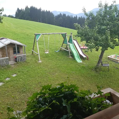 Ein Spielplatz im Grünen mit einer Schaukel, einer Rutsche und einem kleinen Spielhaus. Im Hintergrund sind Bäume und sanfte Hügel zu sehen.