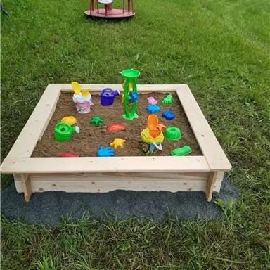 Ein Sandkasten aus Holz mit buntem Spielzeug ist auf einer grünen Wiese zu sehen. Im Hintergrund steht ein Spielgerät.