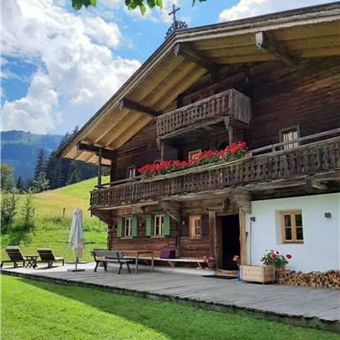A charming wooden house with balconies and flower boxes. It is situated in a green landscape under a clear blue sky.