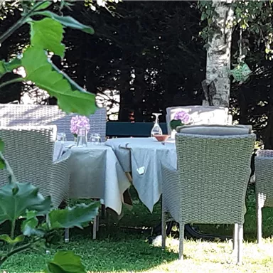 Ein schöner Tisch mit Stühlen im Garten, gedeckt mit einer Tischdecke. Im Hintergrund sind Bäume und grünes Gras zu sehen.