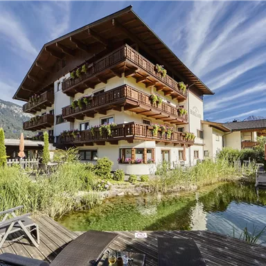 Ein hübsches Gebäude im alpinen Stil mit Balkonen und vielen Pflanzen. Umgeben von Natur und einem kleinen Teich, strahlt es eine ruhige Atmosphäre aus.