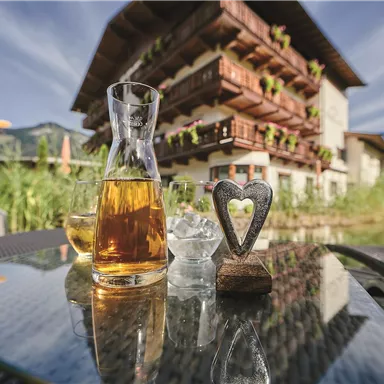 Ein Getränke-Set mit einer Karaffe und Gläsern steht auf einem Tisch. Im Hintergrund ist ein traditionelles Haus mit Balkonen und blühenden Pflanzen zu sehen.