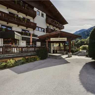 Ein gemütliches Hotel mit traditioneller Architektur in einer Berglandschaft. Der Eingang ist von grünen Pflanzen und einem klaren blauen Himmel umgeben.