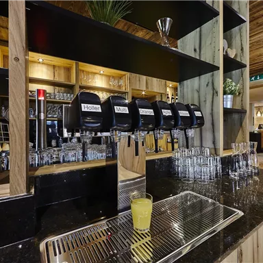 Eine stilvolle Bar mit Holzregalen und modernen Getränkespendern. Im Vordergrund befindet sich ein Glas mit gelbem Getränk auf der Theke.