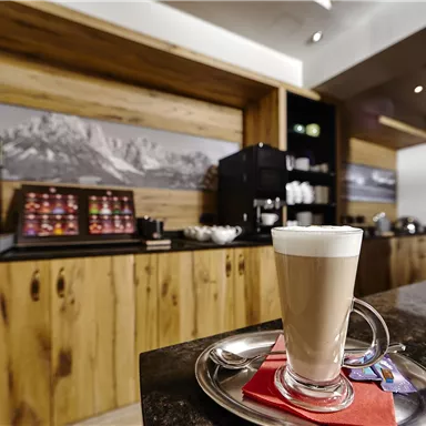 Ein Glas Latte Macchiato steht auf einem Tisch neben einer eleganten Holzoberfläche. Im Hintergrund sind eine Kaffeemaschine und verschiedene Getränke angeordnet.