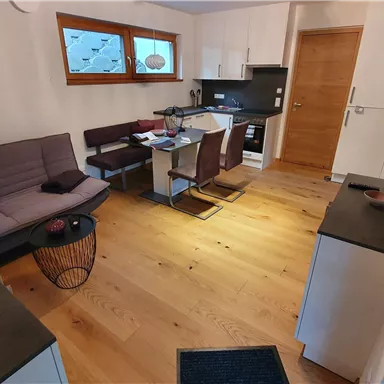 A modern living room with a gray sofa and a dining area. The open kitchen is well-equipped and the floor is made of light wood.