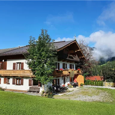 Ein traditionelles alpenländisches Haus mit einem Balkon und bunten Blumen. Die Umgebung ist grün mit Bergen und Wolken im Hintergrund.