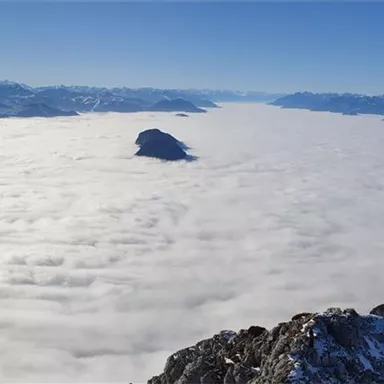 A breathtaking view of snow-covered mountains rising above a dense blanket of clouds. The sky is clear and blue, making the landscape appear even more majestic.