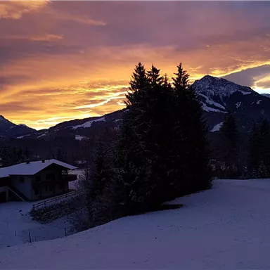 Eine malerische Winterlandschaft mit schneebedeckten Bergen und einem beeindruckenden Sonnenuntergang. Im Vordergrund steht ein kleines Haus, umgeben von Bäumen.