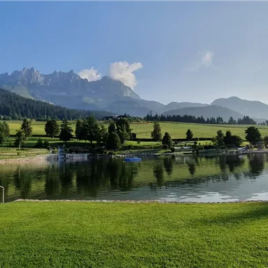 Ein malerischer See umgeben von grünen Wiesen und Bergen. Der Himmel ist klar und die Landschaft strahlt Ruhe aus.