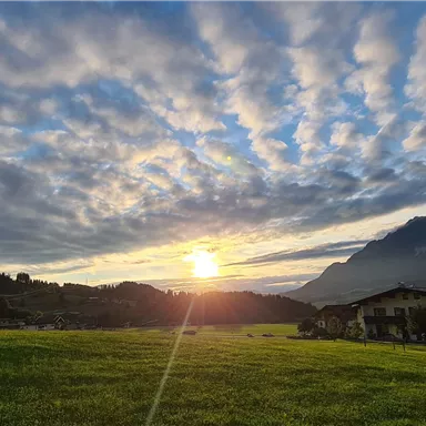 Ein malerischer Sonnenuntergang über einem grünen Feld, umgeben von Bergen. Der Himmel ist mit aufgelockerten Wolken in sanften Farben gefüllt.