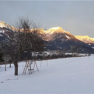 Eine winterliche Landschaft mit schneebedeckten Feldern und majestätischen Bergen im Hintergrund. Ein Baum und eine Schaukel sind im Vordergrund sichtbar.