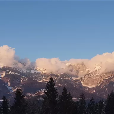 Ein majestätisches Gebirge mit schneebedeckten Spitzen und Wolken. Der klare Himmel und die umliegenden Bäume schaffen eine ruhige Atmosphäre.