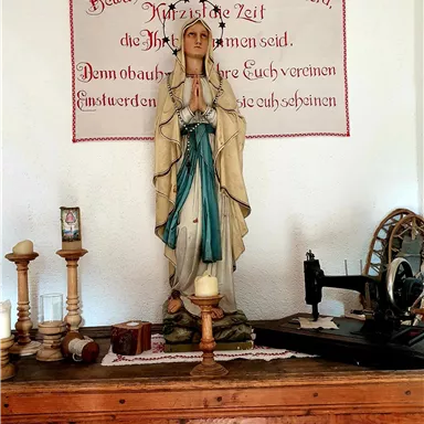 A statue of the Virgin Mary stands on a wooden dresser. In the background, a banner with a religious saying hangs.
